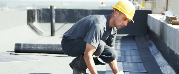 A construction worker in a yellow hard hat installs waterproofing material on a flat rooftop under bright daylight, showcasing expert Residential Roofing skills.