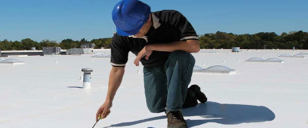 A person wearing a blue hard hat and black shirt is kneeling on a white flat rooftop in North Texas, working with a small tool under a clear sky, with trees and rooftop vents visible in the background—perfect for residential roofing repair.