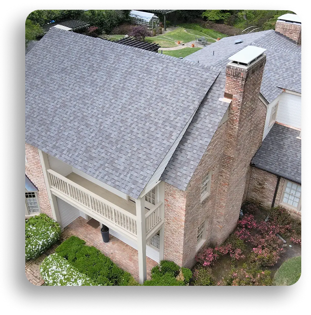 Aerial view of a two-story brick house with a gray shingle roof, showcasing expert residential roofing in North Texas, plus a covered balcony and landscaped yard with green bushes and flowering plants.
