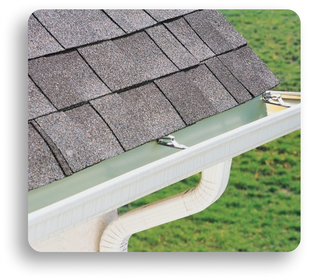Close-up of a house roof corner with gray asphalt shingles and a clean, white rain gutter. Installed by American Patriot Roofing, this residential roofing feature includes a mesh guard to block debris. Green grass is visible in the background.