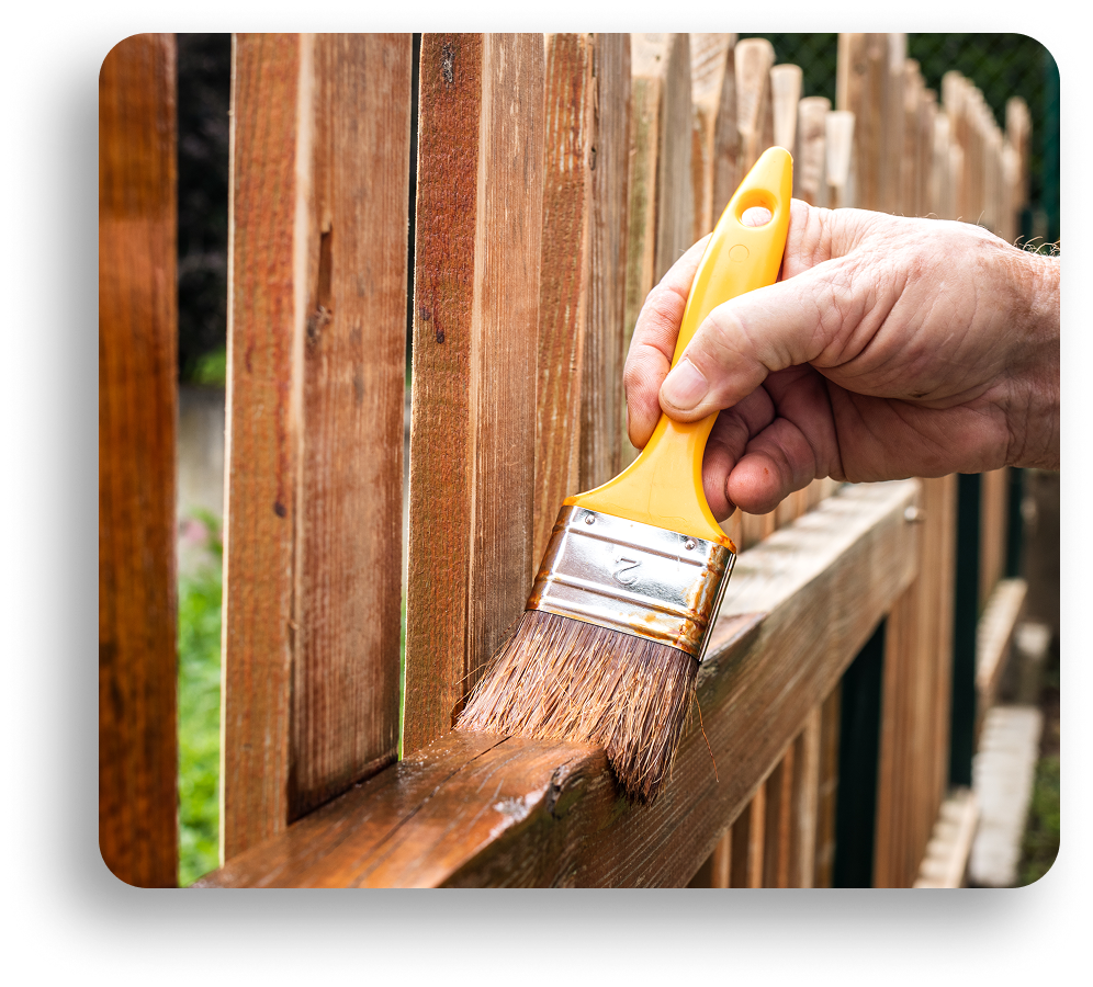 A hand holds a yellow paintbrush, applying brown stain to a wooden fence outdoors in North Texas. The freshly coated slats stand out with a crisp finish, while the background is slightly blurred.