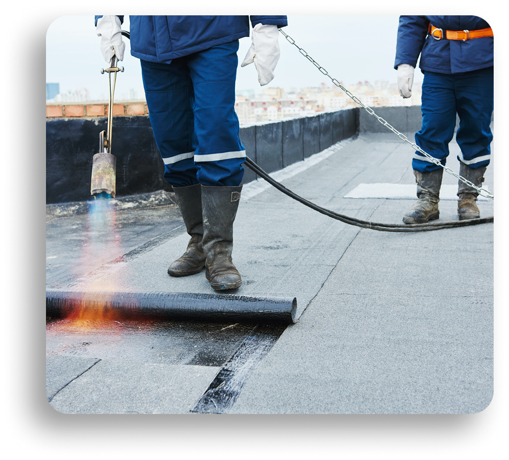 Two workers in blue uniforms and boots install roofing material using a torch, applying heat to secure a black roll onto a flat rooftop. Flames are visible near the roll as American Patriot Roofing expertly handles this commercial roofing repair.