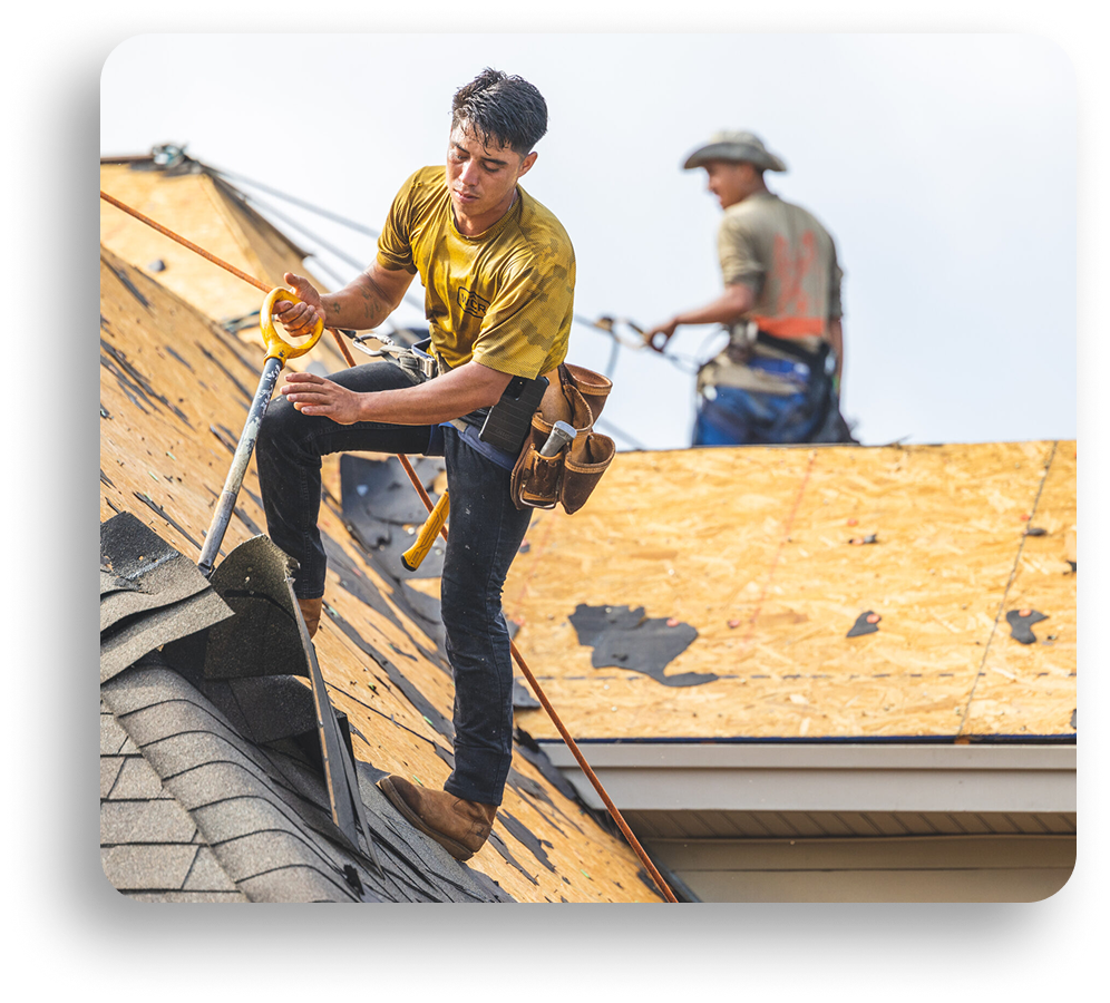 Two construction workers from American Patriot Roofing install shingles on a North Texas roof. One, wearing a safety harness and tool belt, works near the edge, while the other stands in the background on the unfinished wooden roof.