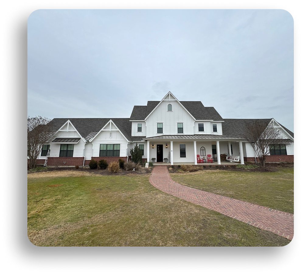 A large white two-story house with dark roof shingles and brick accents, a wraparound porch with chairs, and a curved brick walkway across a grassy lawn—showcasing quality residential roofing by American Patriot Roofing under a cloudy sky.