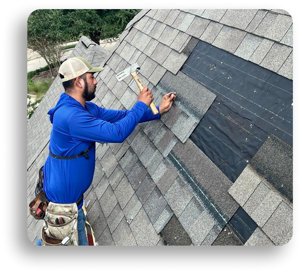 A construction worker wearing a blue shirt, tan cap, and tool belt is repairing a shingled roof in North Texas, holding a nail and hammer while attaching gray roofing shingles for residential roofing.