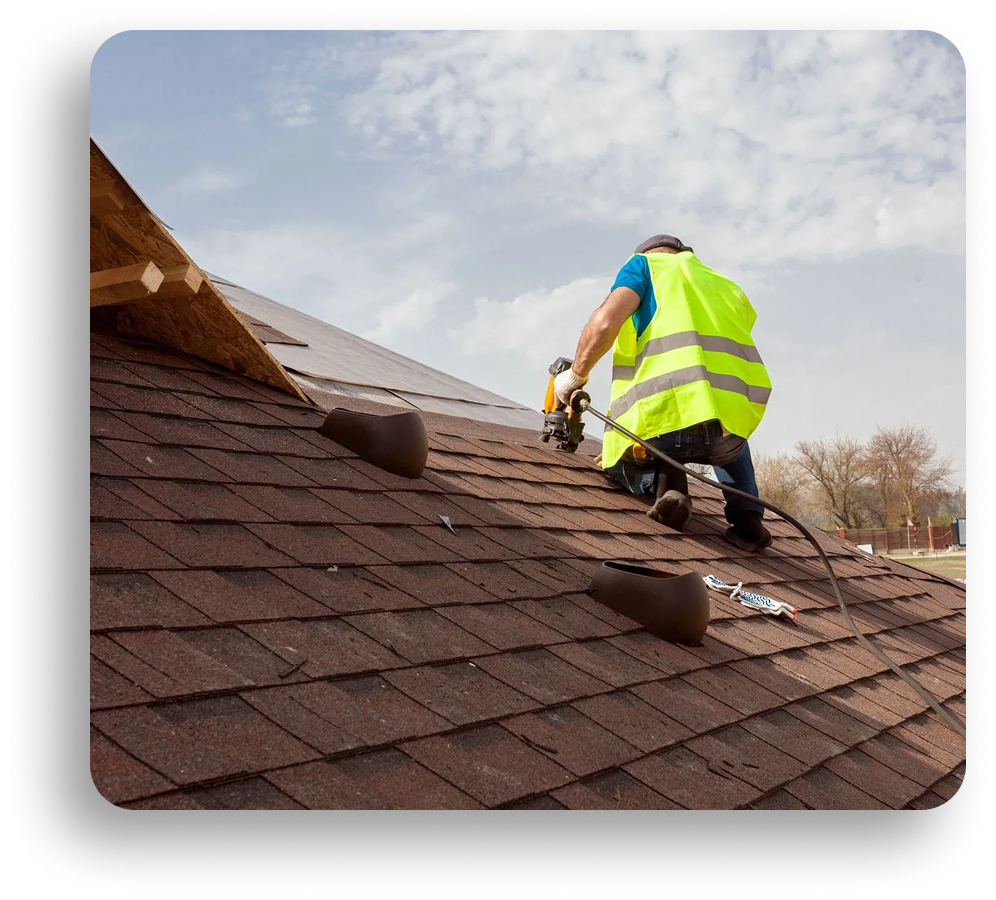 A worker in a neon safety vest uses a nail gun while installing shingles on a sloped roof under a partly cloudy sky, with roofing tools and materials from American Patriot Roofing nearby, specializing in commercial roofing and repairs.