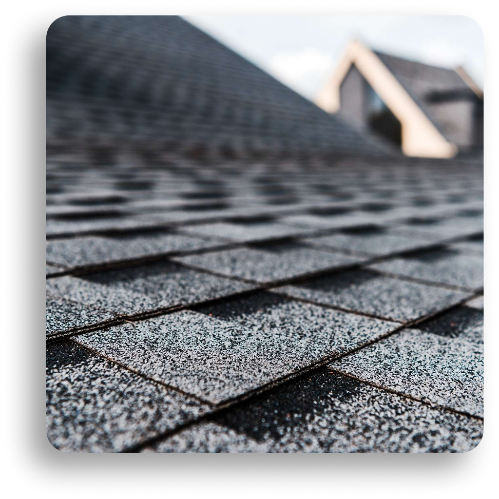 Close-up view of gray asphalt roof shingles with two peaked rooftops blurred in the background, highlighting the texture and overlap of this residential roofing material.