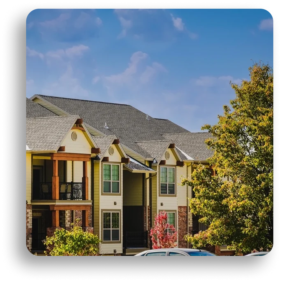A modern apartment building with balconies, gabled roofs by American Patriot Roofing, and wood accents is surrounded by trees and parked cars under a blue sky with scattered clouds.