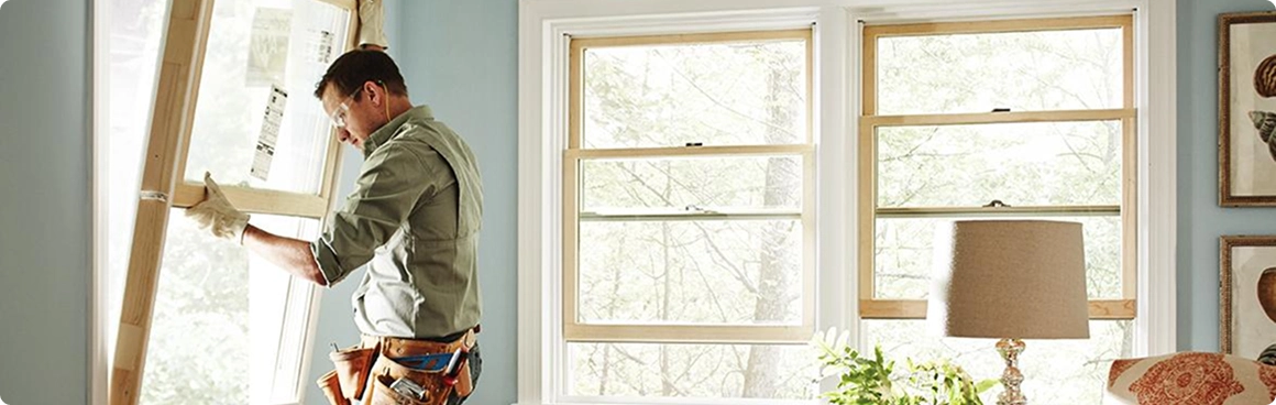 A person wearing work gloves and a tool belt installs a window frame in a brightly lit North Texas room with light blue walls, next to two large windows, a lamp, and a plant—quality work from American Patriot Roofing.