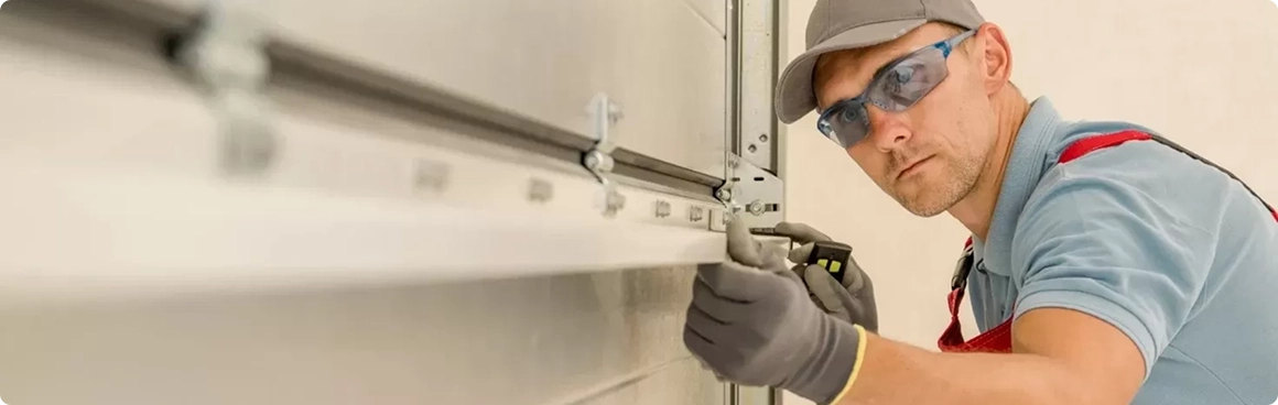 A person wearing safety glasses, gloves, and a cap is using tools to repair or install a metal garage door in North Texas, focusing intently on the work.