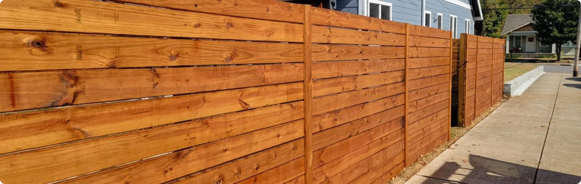 A modern wooden fence with horizontal planks runs alongside a sidewalk in front of a blue house in North Texas, with a tree and other houses visible in the background.