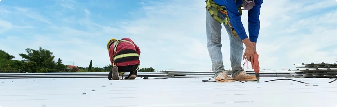 Two workers from American Patriot Roofing, wearing safety harnesses, are installing or repairing a white metal roof under a blue sky. One crouches while the other uses a power tool, with greenery in the background—a scene of expert roofing repair.