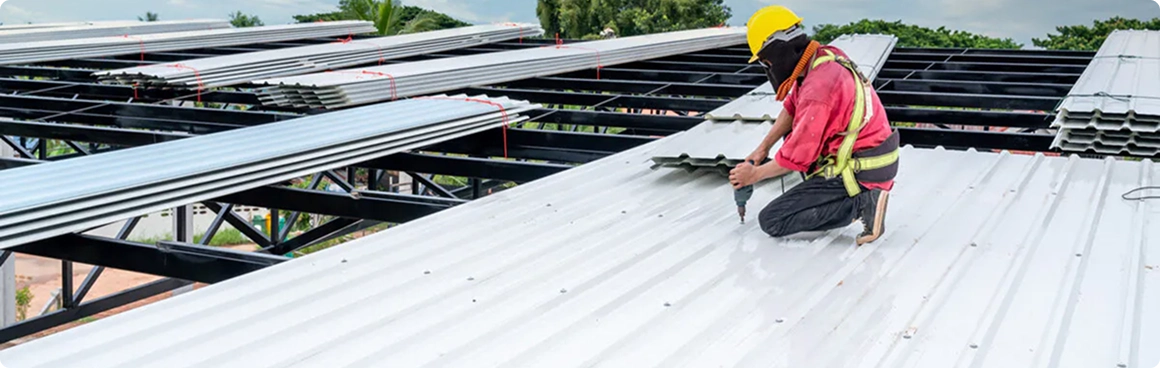 A construction worker in safety gear installs metal roofing sheets on a large building structure for American Patriot Roofing, surrounded by stacks of additional sheets and green trees in North Texas.