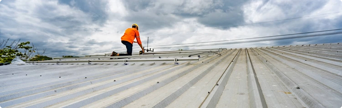 A worker in an orange safety vest and helmet is installing or repairing a metal roof under a cloudy sky, showcasing expert roofing repair for residential roofing in North Texas. The low angle highlights the vast expanse of the corrugated roof.