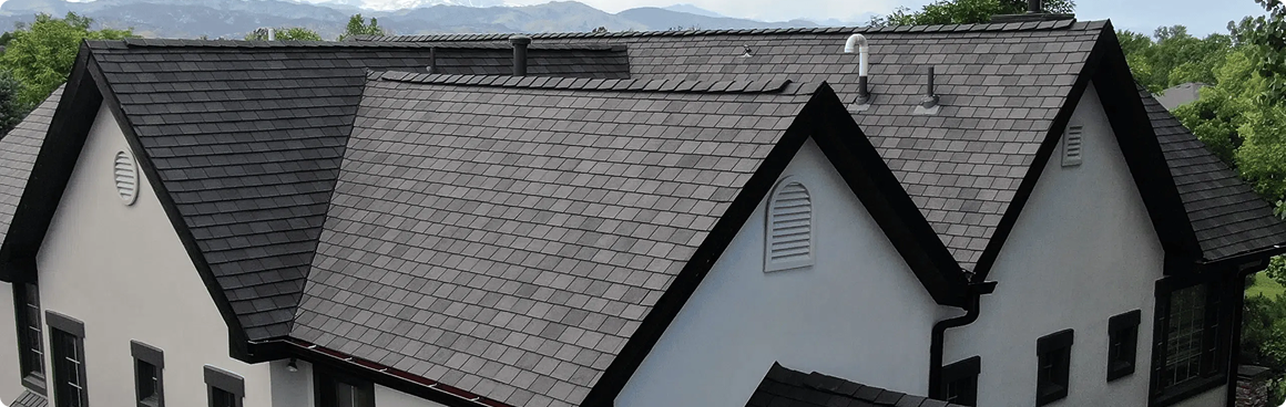 A modern white house with multiple steep gables and dark gray shingle roofing by American Patriot Roofing, nestled among green trees in North Texas, with distant mountains visible in the background.