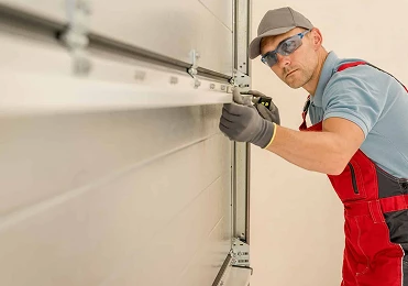 A worker in red overalls, a light blue shirt, gloves, safety glasses, and a cap is installing or repairing a garage door for American Patriot Roofing in North Texas, using a tool to adjust the metal track.