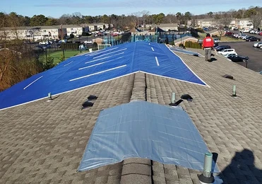 Workers install blue and gray tarps on a shingled roof, likely for weather protection or repairs—a typical scene in residential roofing, with parked cars and a neighborhood visible in the background.