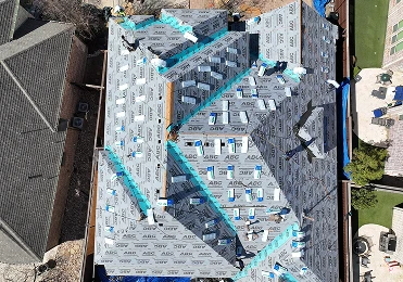 Aerial view of a house roof under construction in North Texas, covered with synthetic underlayment and blue tape. Workers from American Patriot Roofing install materials, with nearby yards and structures partially visible.
