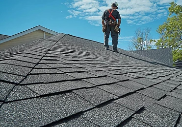 A worker from American Patriot Roofing, wearing a tool belt and safety harness, stands on a gray shingled roof under a blue North Texas sky with scattered clouds, inspecting or performing roofing repair.