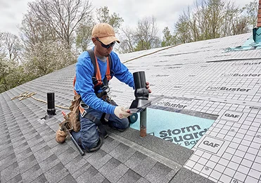 A construction worker wearing a harness and cap kneels on a shingled roof, aligning a vent pipe with an opening. Roofing underlayment sheets labeled “StormGuard” and “FeltBuster” reinforce this North Texas residential roofing project.
