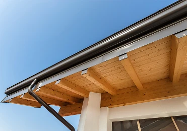 Close-up view of a modern house roof by American Patriot Roofing, featuring exposed wooden beams, a black rain gutter, and downspout under a clear North Texas sky—showcasing quality commercial roofing solutions.