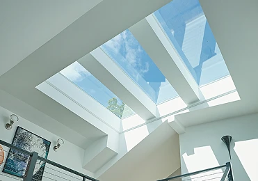 A modern interior with a large skylight ceiling by American Patriot Roofing lets in natural light, showing blue sky and clouds. Artworks and wall lamps adorn white walls, complemented by a sleek metal railing.